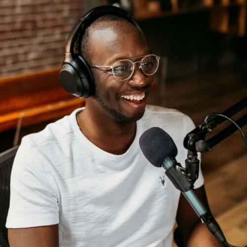 Bald and slim black men wearing glasses, black headphones, and a white tee is smiling at the computer and has a microphone in fron of him.